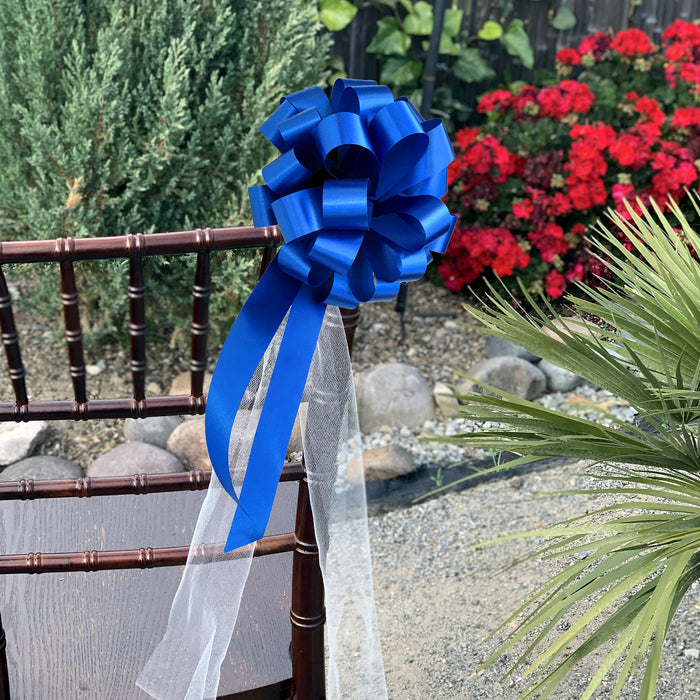 A large Santorini blue ribbon bow with long tails and a sheer white ribbon, tied to the back of a brown wooden chair in an outdoor garden setting with red flowers and greenery.