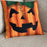 A square decorative throw pillow featuring a carved jack-o'-lantern face design in orange, black, and green, resting on a textured gray fabric sofa.